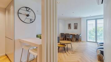 Un salon moderne avec un parquet en bois disposé en chevrons, un coin repas comprenant une table et des chaises en bois, et une grande horloge murale.