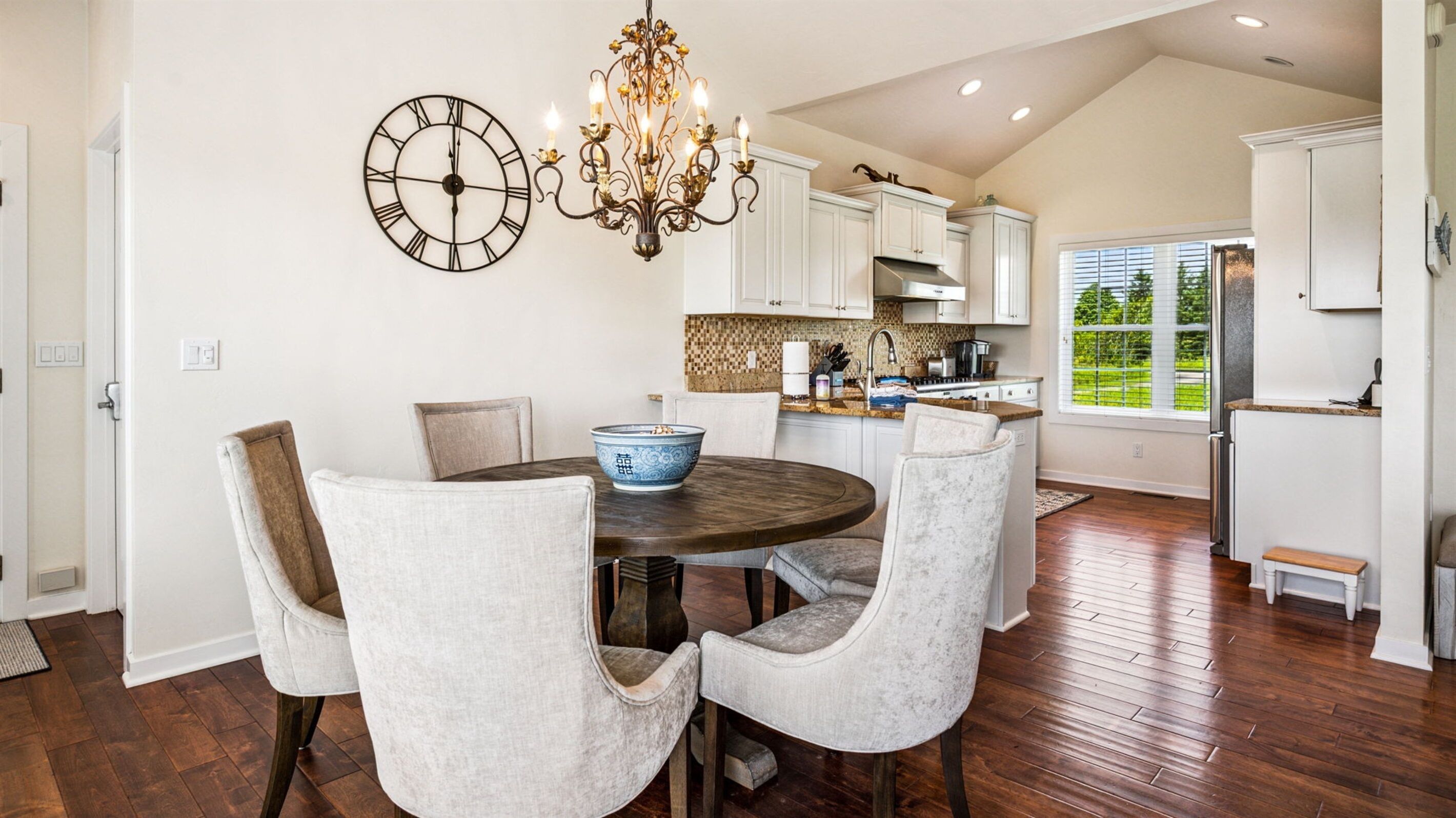 A dining area with a round table, four chairs, a wall clock, and a chandelier.