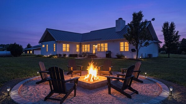 A house with a fire pit in the center surrounded by chairs, a well-maintained lawn, and a clear evening sky.