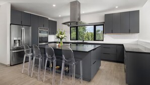 A modern kitchen with a central island, grey cabinetry, stainless steel appliances, and a large window providing a view of greenery. - Villa Eden, Modern Oceanfront Villa on Blue Mountain (Providenciales)