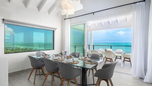 A dining area with a glass table, grey chairs, and a view of the ocean. - Villa Eden, Modern Oceanfront Villa on Blue Mountain (Providenciales)