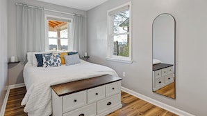 Een slaapkamer met een bed, een dressoir, een raam en een spiegel.
