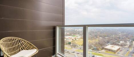 A balcony with a wicker chair, a view of a cityscape, and a brick floor.