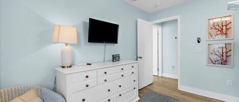 A room with a white dresser, a flat-screen TV, a lamp, and a colorful rug.