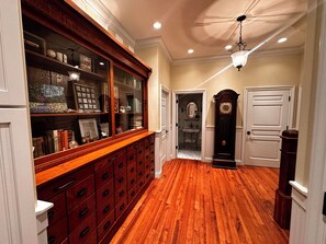 A hallway with wooden flooring, a glass-fronted cabinet with drawers, a grandfather clock, and a door with a mirror.