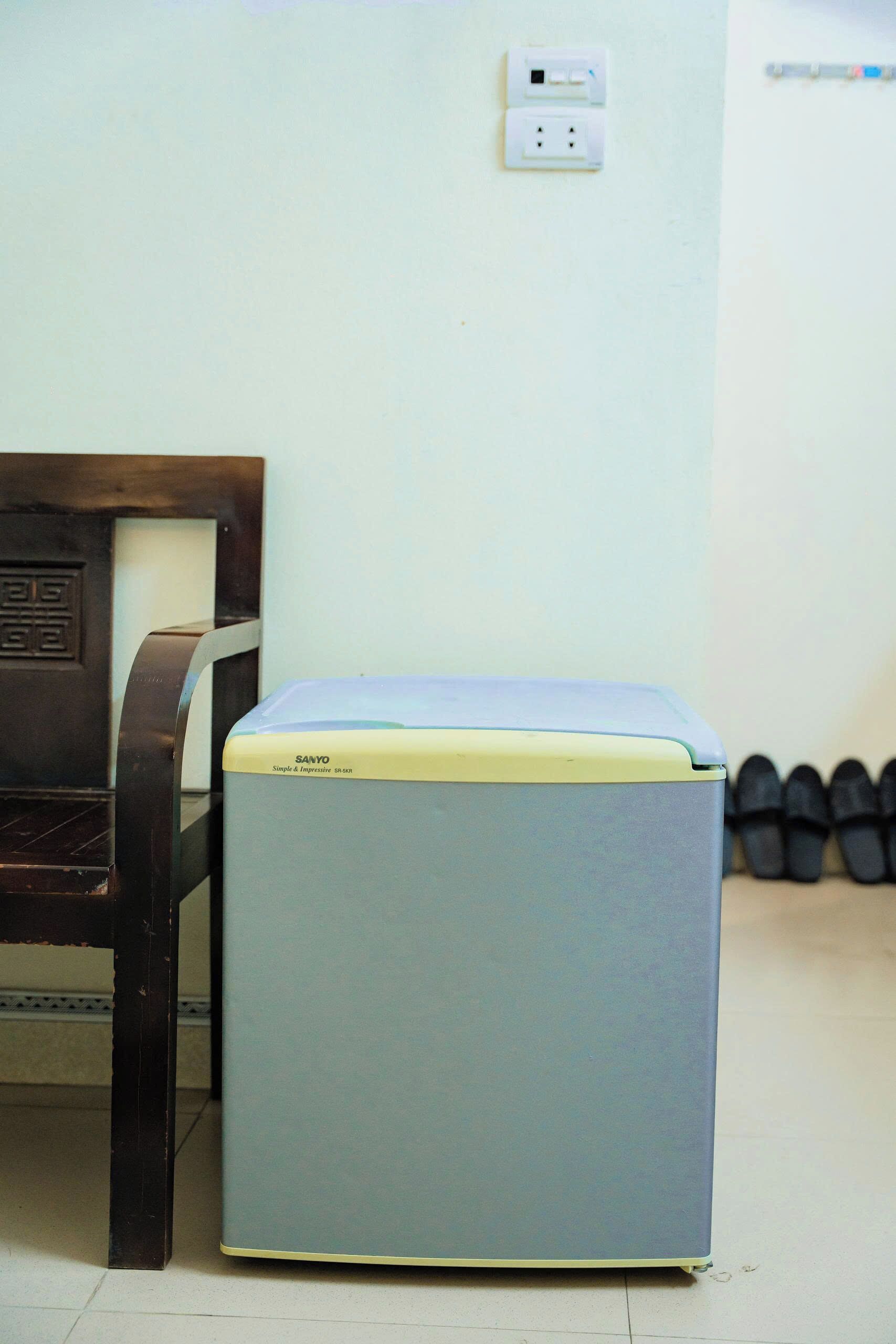 Un refrigerador pequeño azul claro con parte superior amarilla, una silla de madera y un enchufe de pared.
