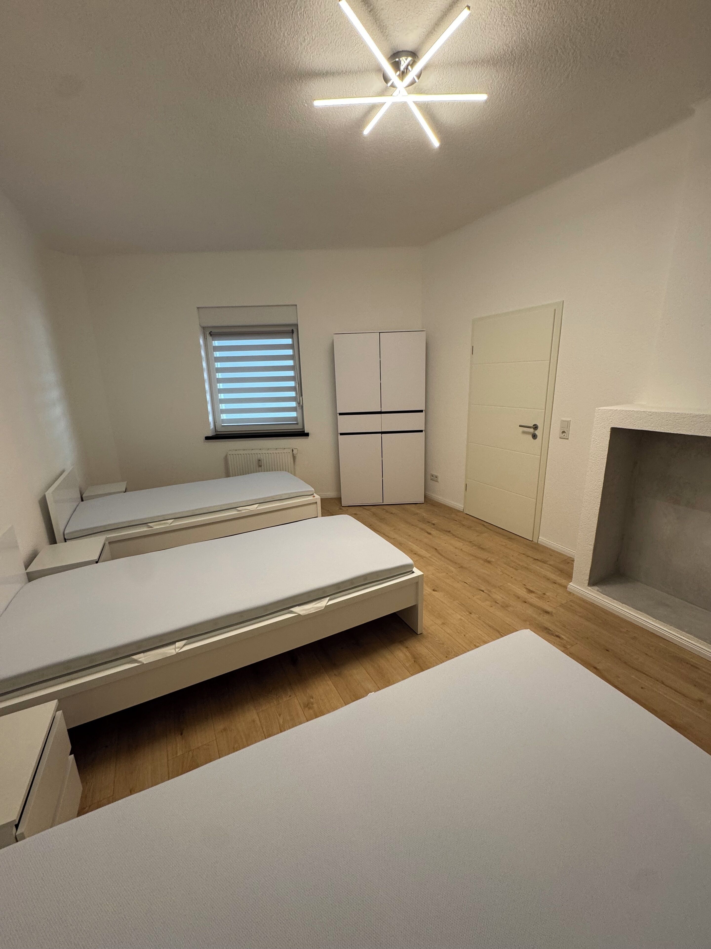 A minimalist bedroom with two beds, a wardrobe, a window with blinds, and a ceiling light fixture.