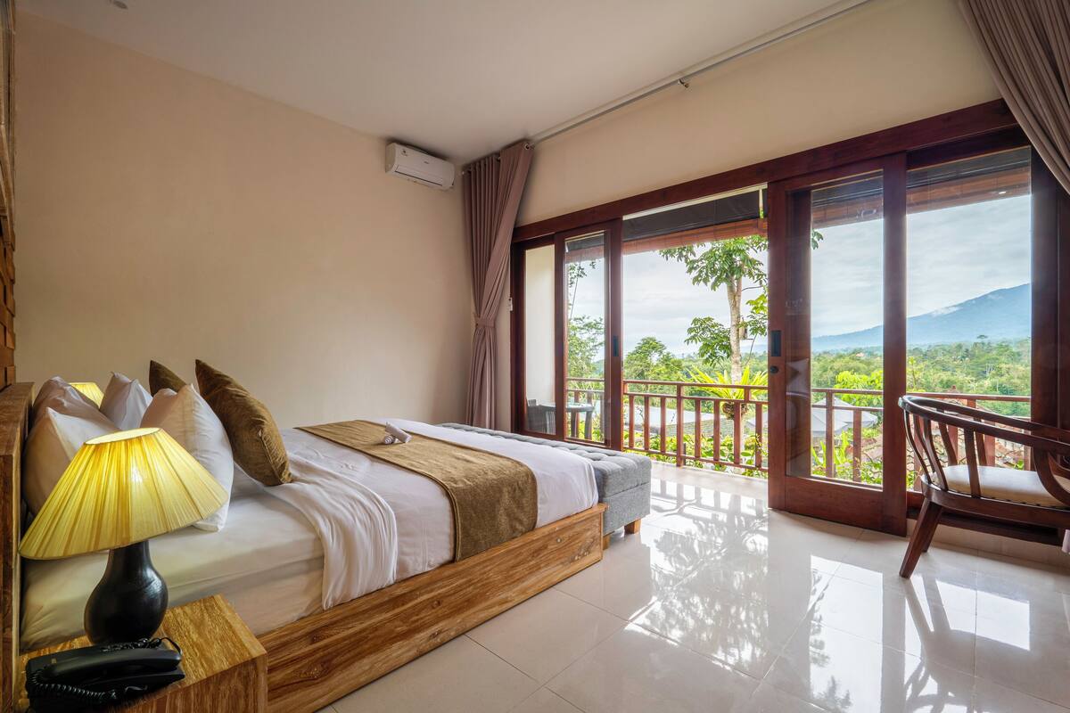 A bedroom with a bed, a bedside lamp, a wooden nightstand, a chair, and a balcony with a view of greenery and mountains.