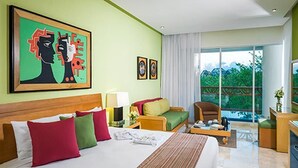 Ein Hotelzimmer mit einem großen Bett, einer Couch, einem Sessel, einem Schreibtisch und einem Balkon mit Blick auf Grünflächen.