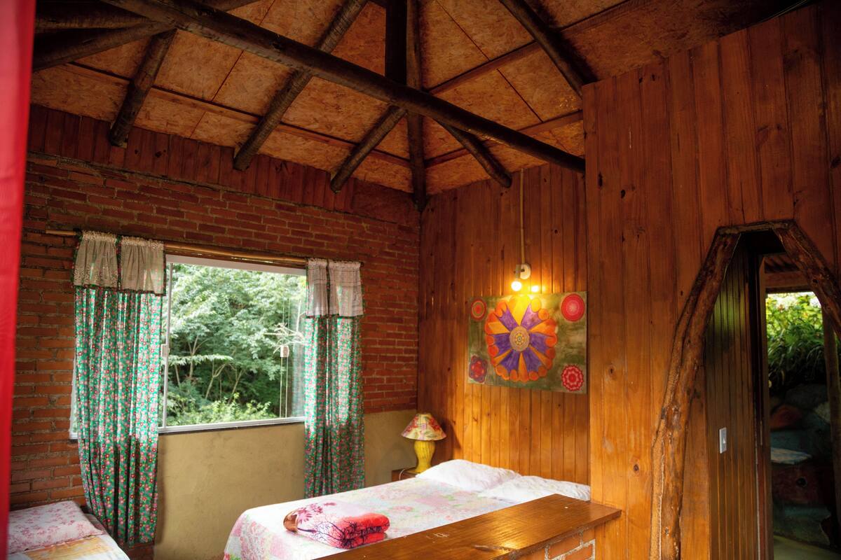 A wooden cabin room with a bed, a window with curtains, a lamp, and a door leading outside.