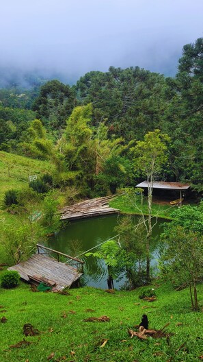 A rustic wooden bridge over a pond, surrounded by lush greenery and a small hut with a corrugated metal roof. - Chalés Nova Friburgo (Nova Friburgo)