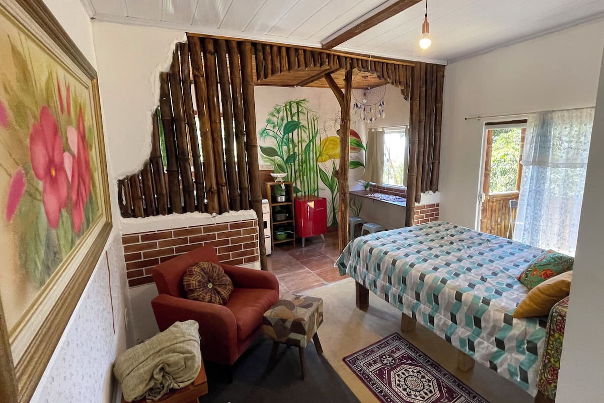 A room with a bed, a red chair, a small table, a decorative wall, and a window with a view of greenery.