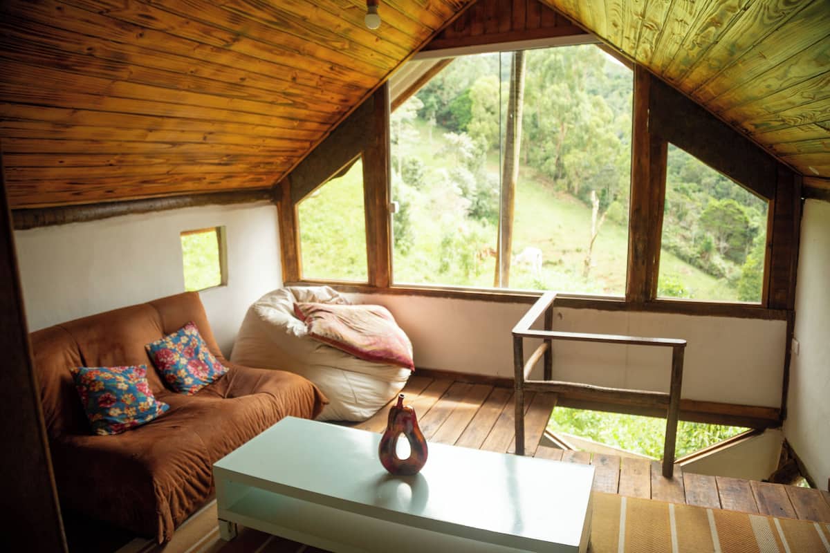 A cozy room with a brown couch, a white table, and a large window offering a view of greenery outside.