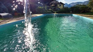 A clear swimming pool with a water fountain, surrounded by lush greenery and a mountain backdrop.