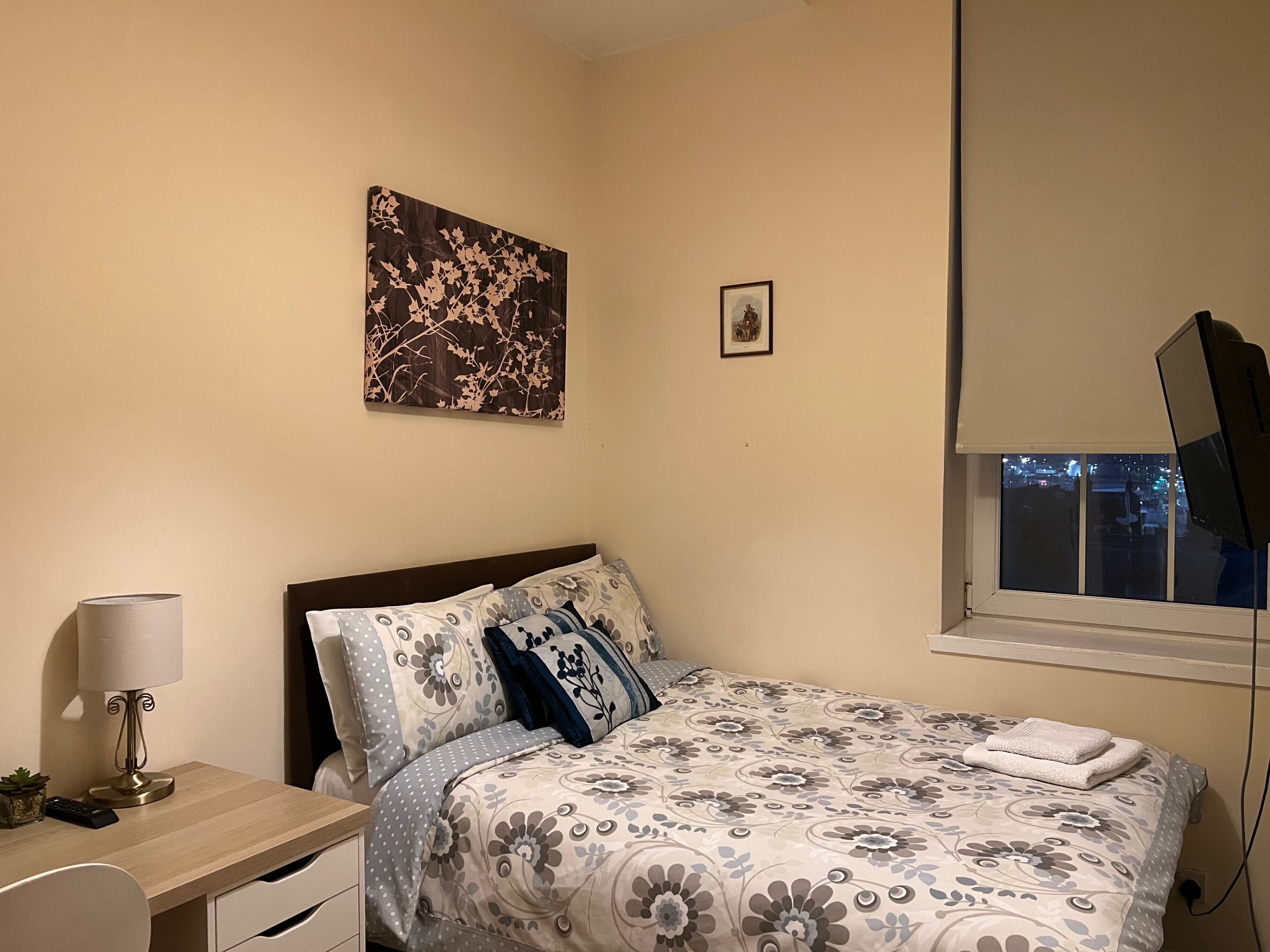 Un dormitorio con cama, escritorio, lámpara, un marco de fotos y ventana con vista a la ciudad.