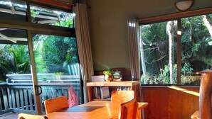 Una mesa de madera con sillas, un pequeño estante con libros y una planta, y vistas al verdor exterior.