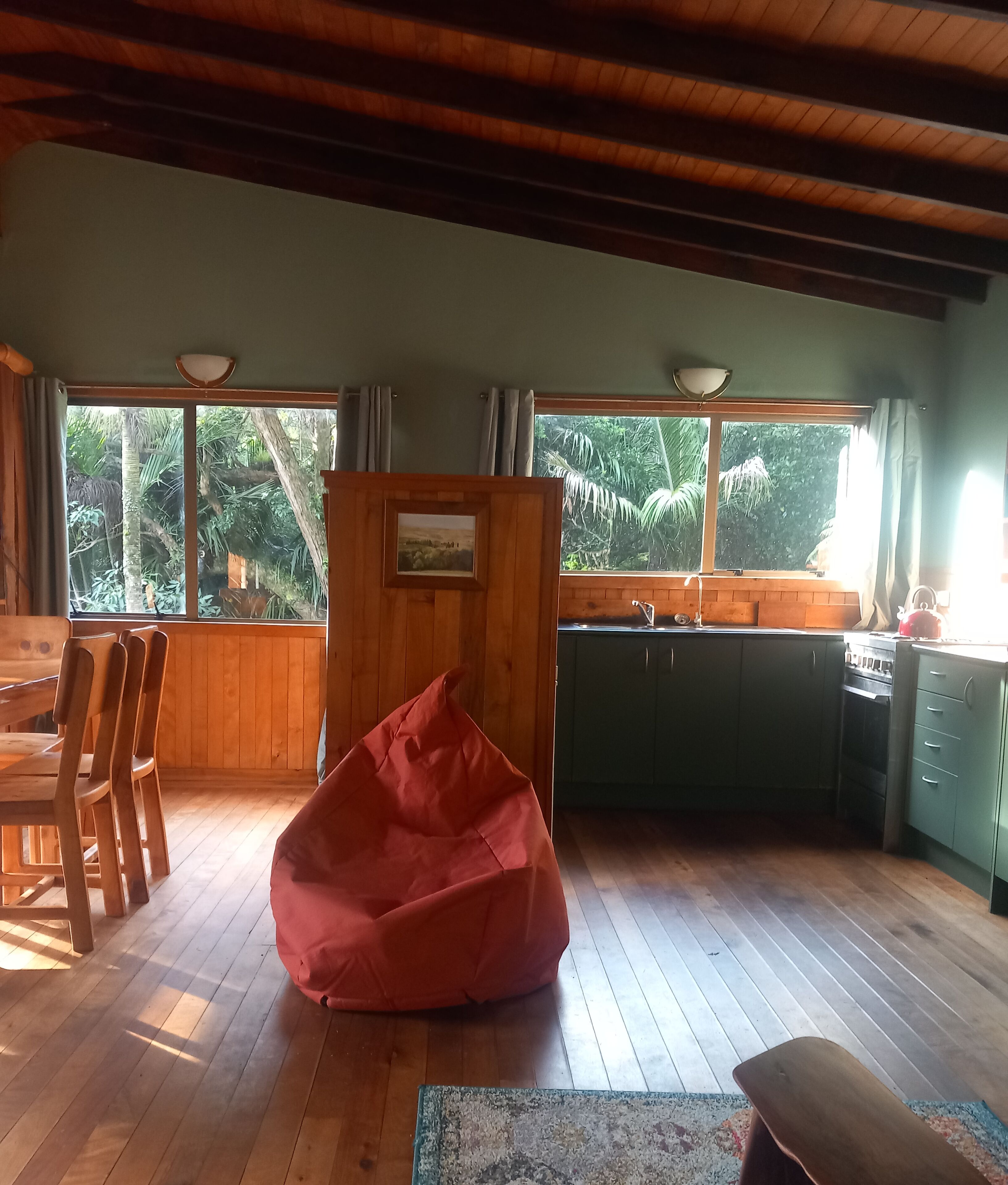 Una stanza con pavimento in legno, un pouf rosso, un tavolo da pranzo con sedie, un angolo cottura e ampie finestre con vista sul verde.