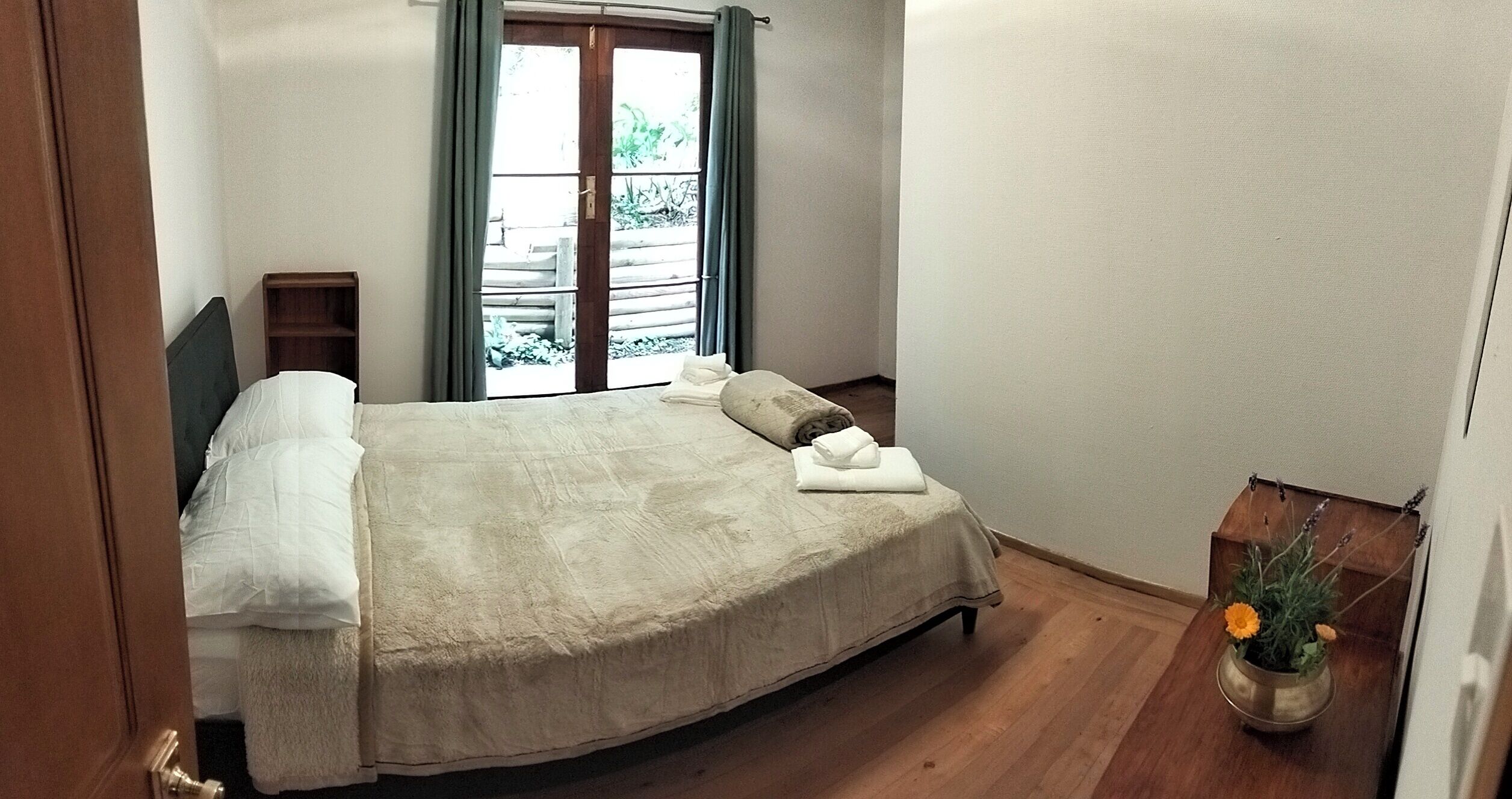Un dormitorio con cama, cabecero de madera, ventana con cortinas, mesita de noche de madera y una planta en maceta.