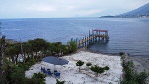 A beach with a wooden pier extending into the water, surrounded by trees and a small seating area with an umbrella.