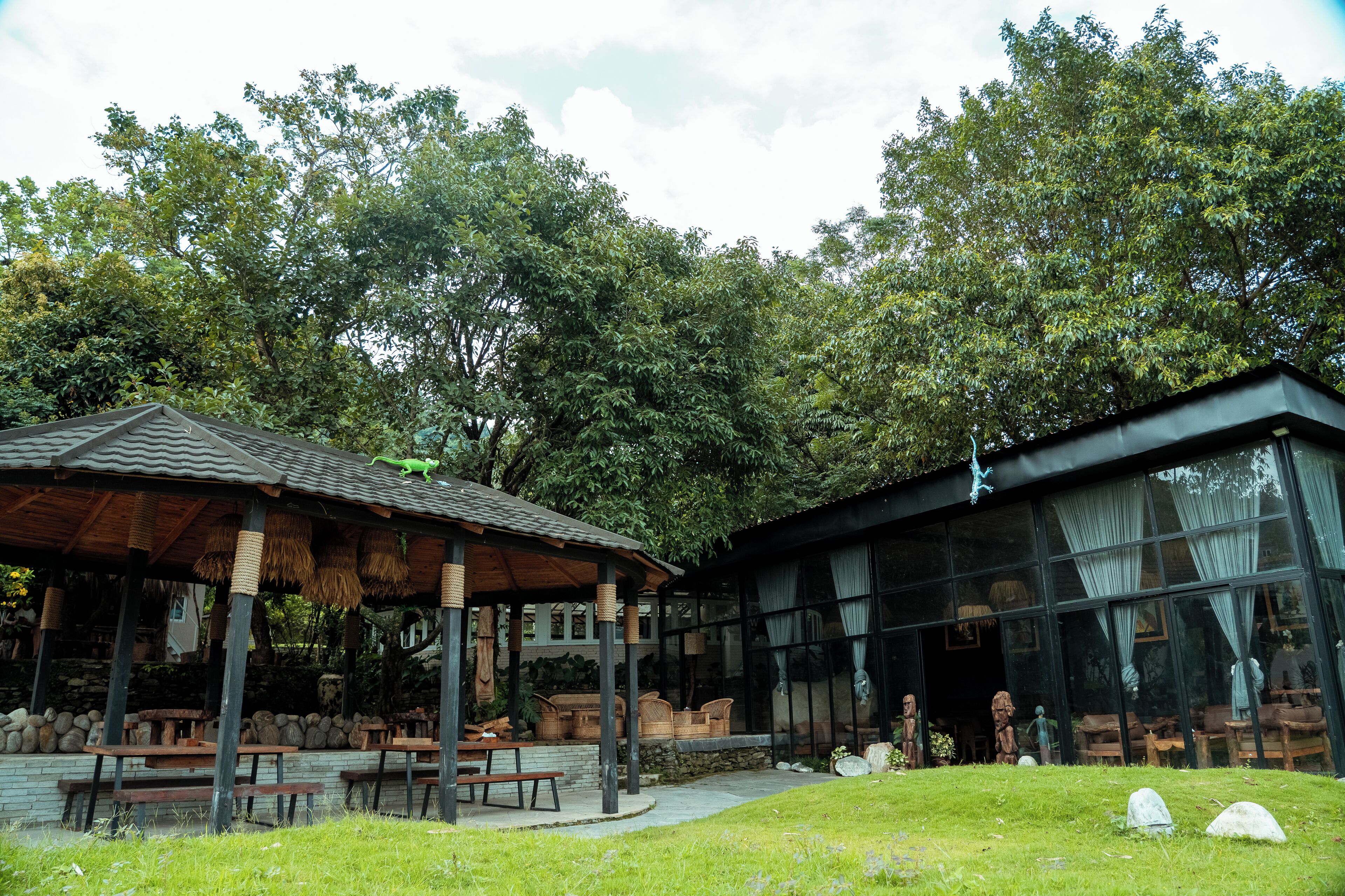 A rustic outdoor seating area with wooden tables and benches, surrounded by lush greenery and a stone wall.