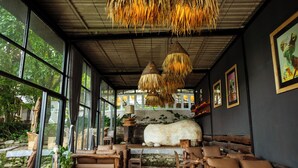 A room with large windows, a stone wall, and a ceiling with hanging thatch. There are wooden furniture pieces and a large white object on the wall. - Krazy Gecko Resort (Pokhara)