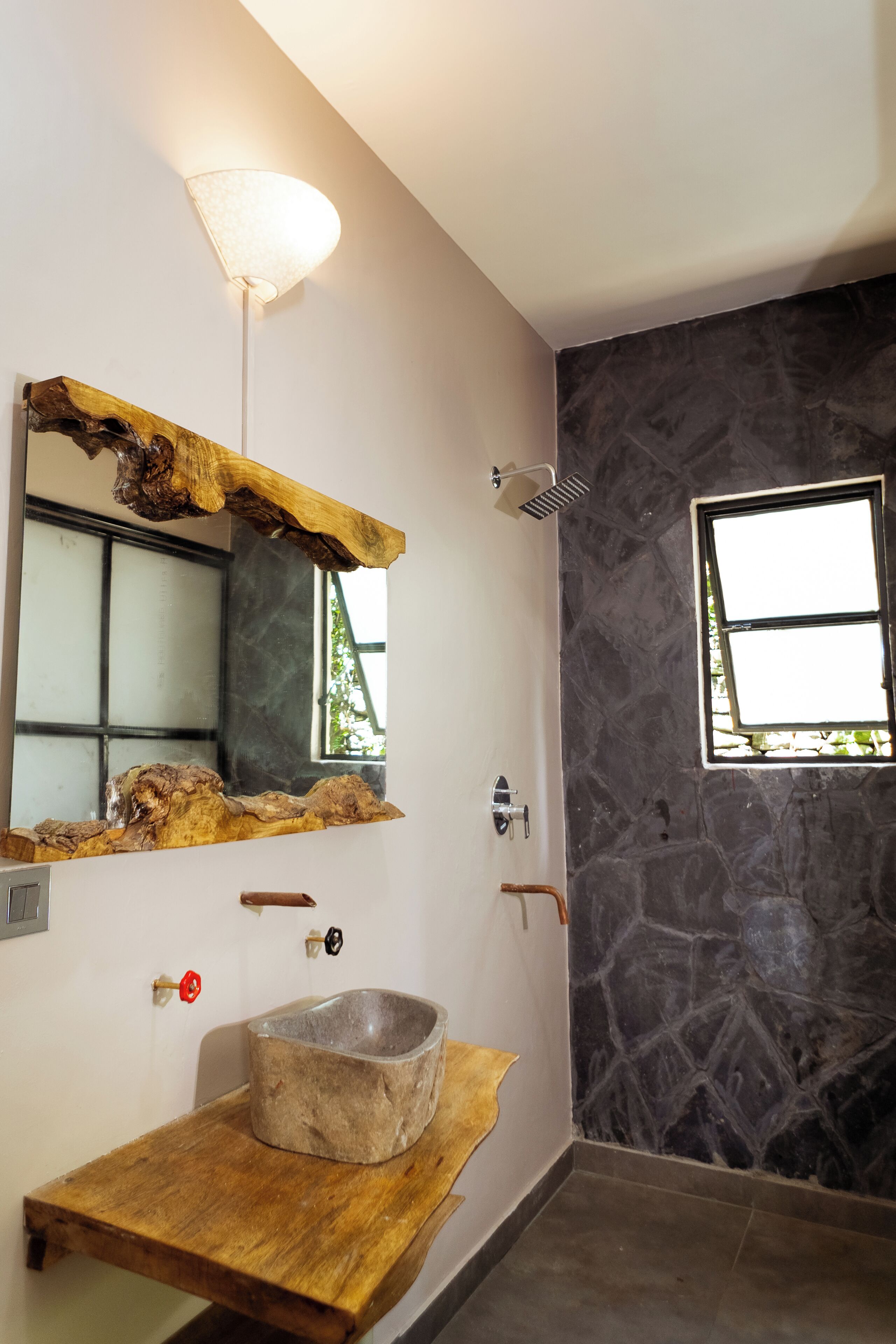 Un baño con pared de piedra, un tocador de madera, un lavamanos, un espejo con marco de madera, una lámpara y una ventana.