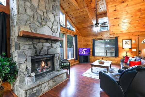 A cozy cabin interior with a stone fireplace, wooden walls, a ceiling fan, and a television.
