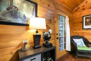 Interior acogedor de una cabaña con pared de madera, una lámpara, una estatua de oso, una silla con cojín y un cuadro enmarco.