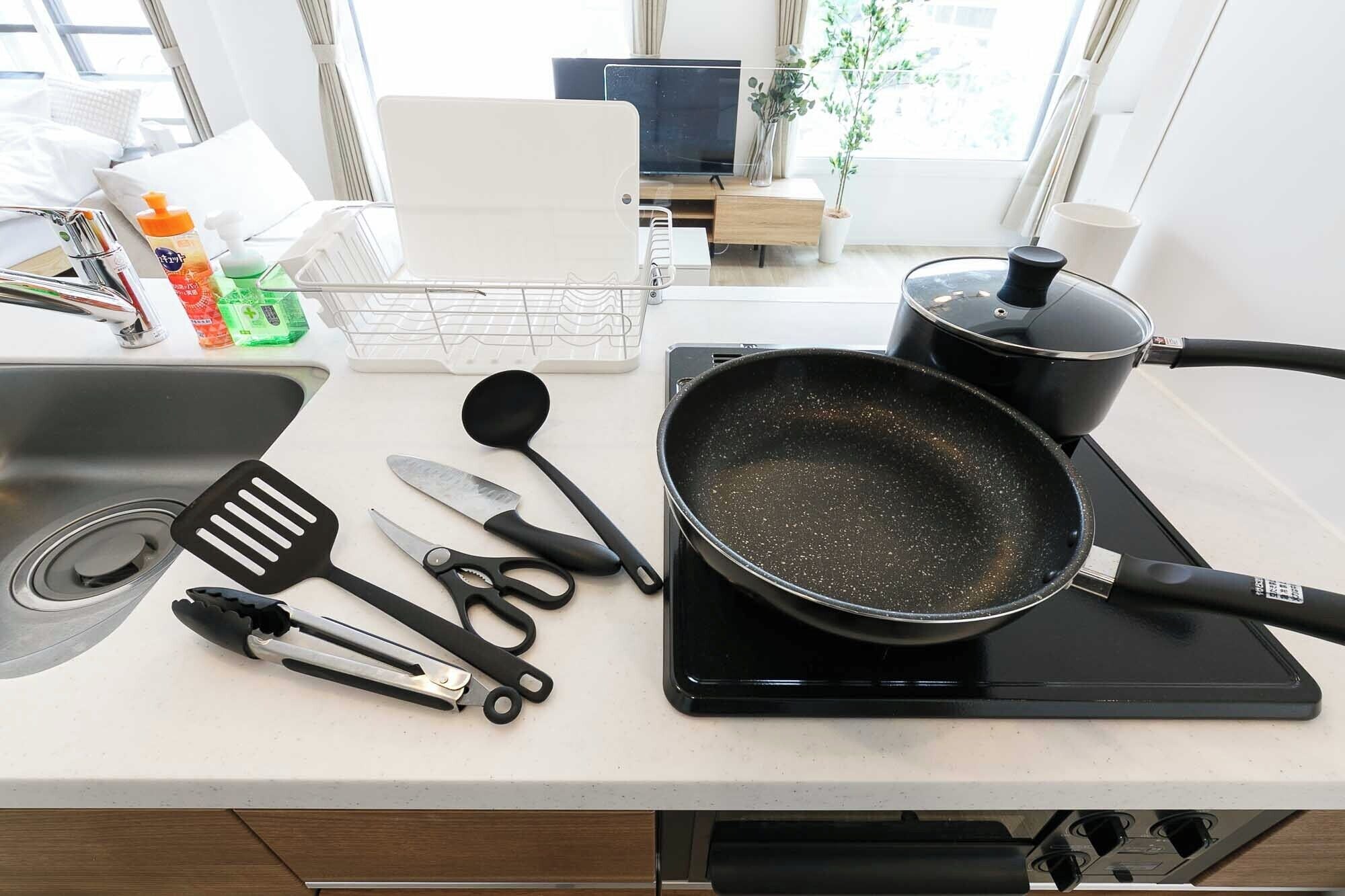 Una encimera de cocina con una sartén sobre una hornilla portátil, diversos utensilios de cocina, un lavamanos y una ventana con vistas al exterior.
