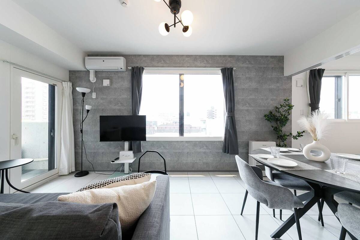 A modern living room with a grey sofa, a dining table with chairs, a flat-screen TV, and a large window with curtains.