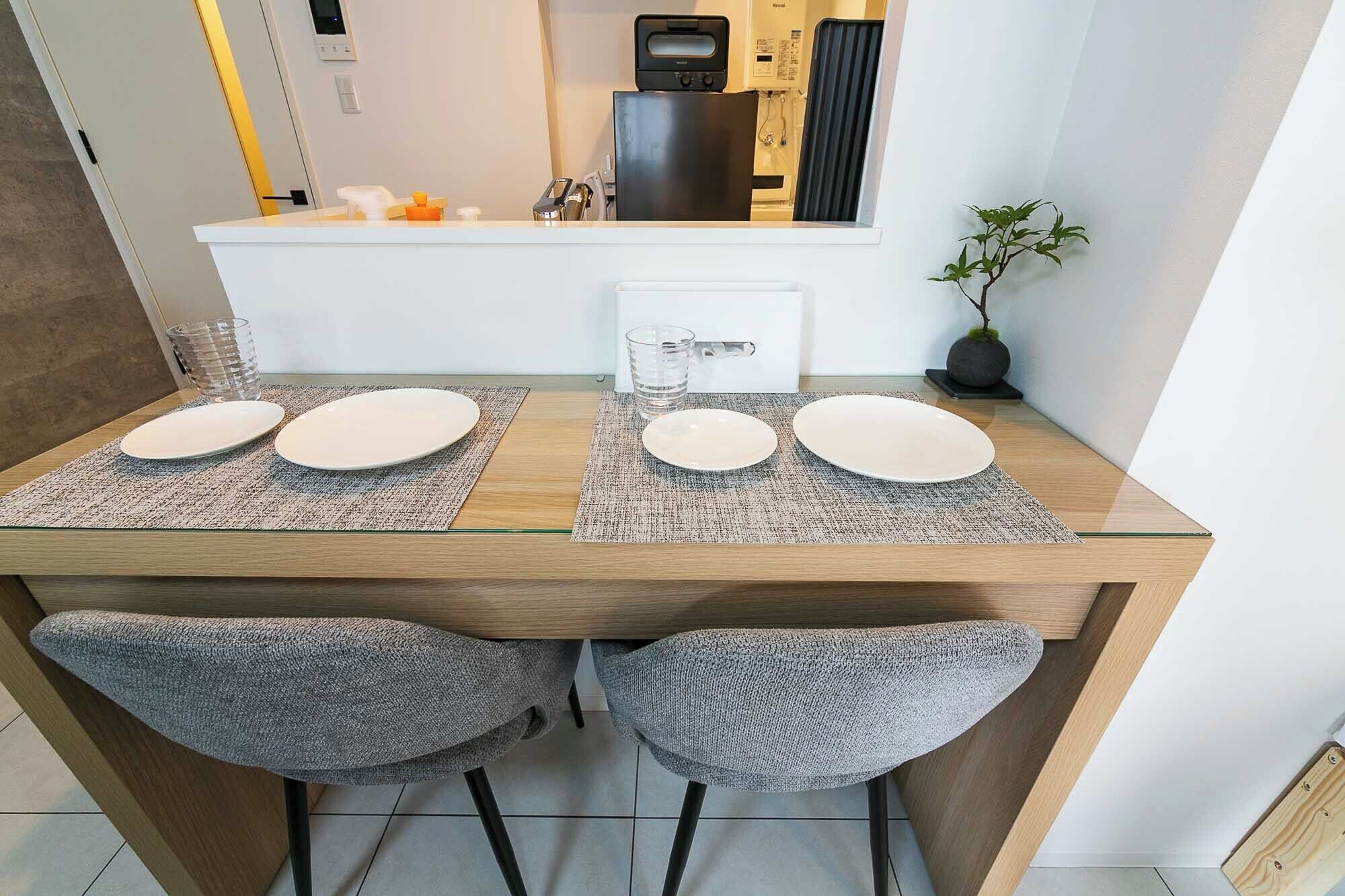 A compact dining area with a wooden table, four white plates, two grey chairs, and a small potted plant.