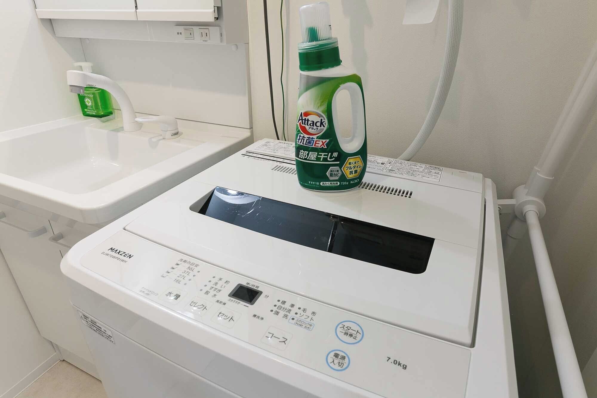 A white washing machine with a control panel, a green bottle of detergent, and a white sink with a soap dispenser.