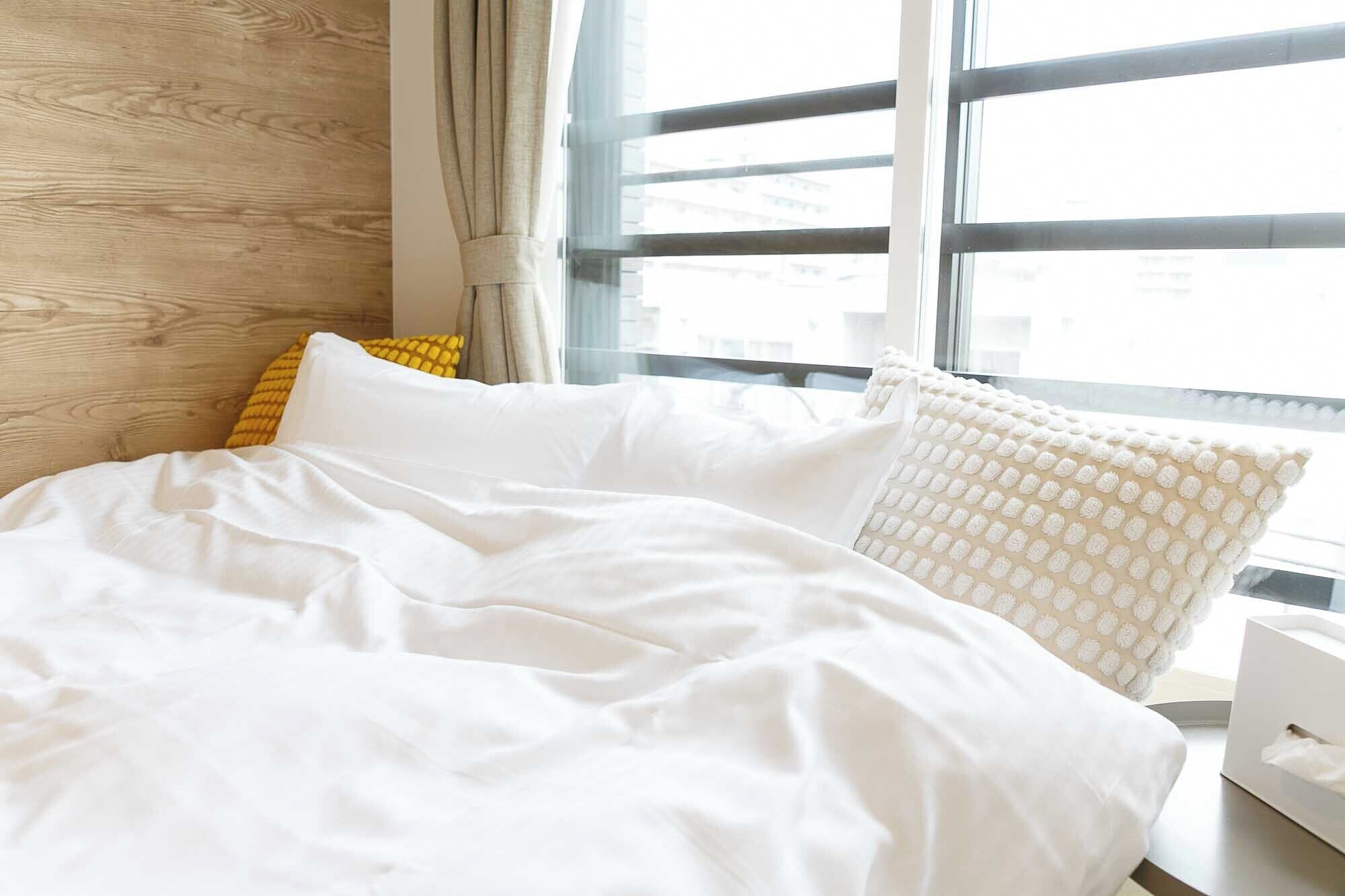 Cama bem arrumada com roupa de cama branca e um travesseiro amarelo, janela com vista para edifícios e cabeceira de madeira.