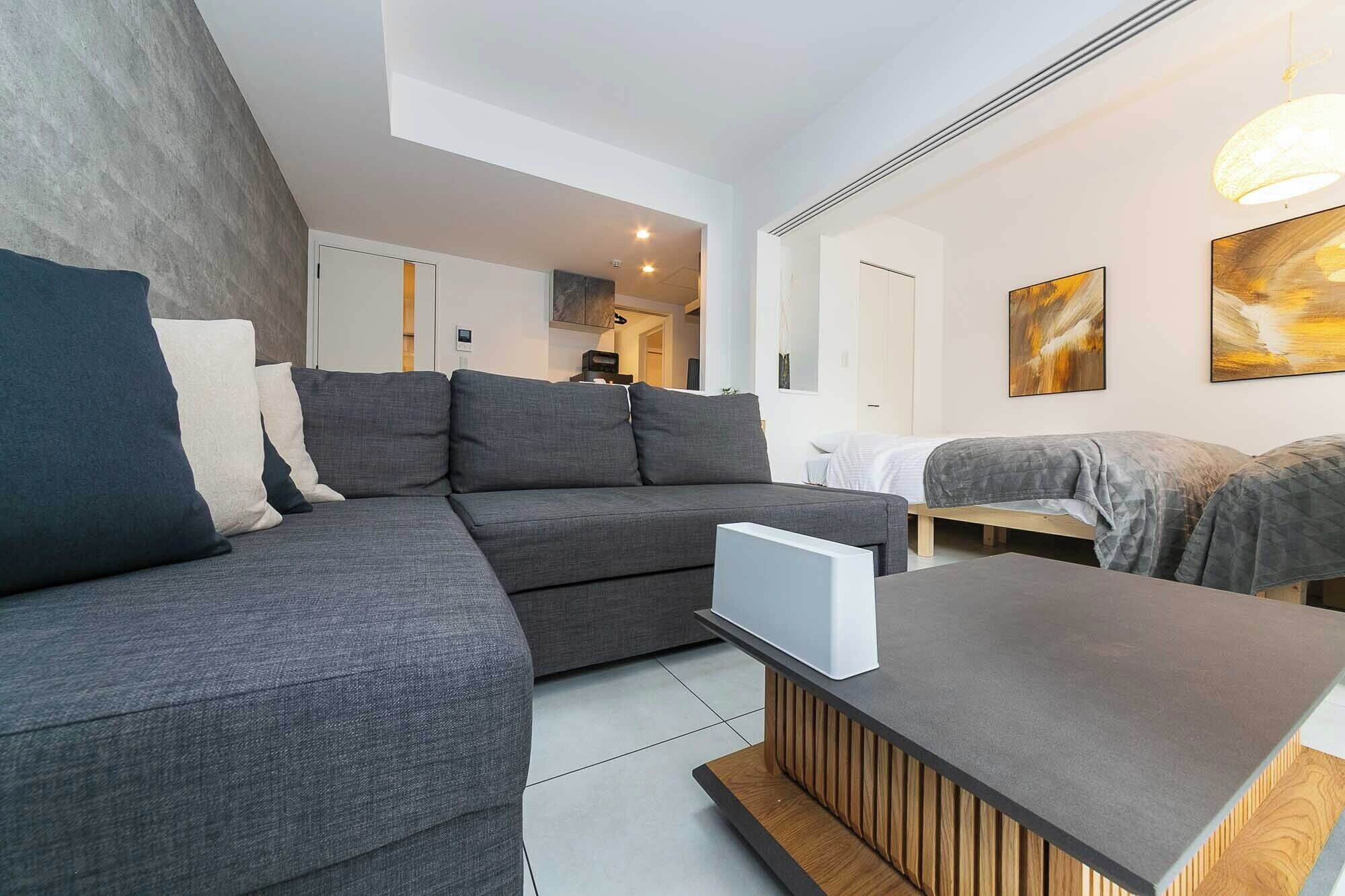 Un salon moderne avec un canapé d’angle gris, une table basse en bois et un lit recouvert d’une couverture grise. Des tableaux abstraits ornent le mur, et un luminaire est fixé au plafond.