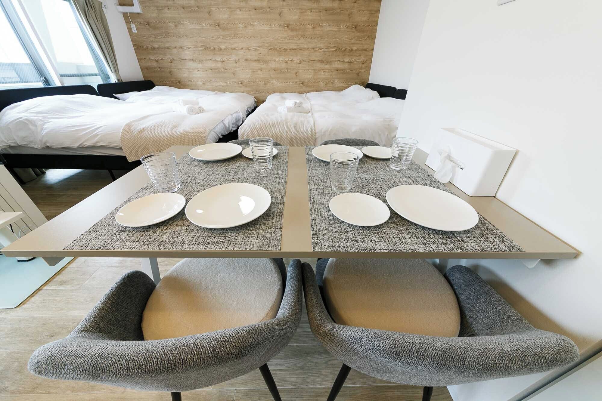A modern hotel room with a bed, a table set for two with plates and glasses, and two upholstered chairs.