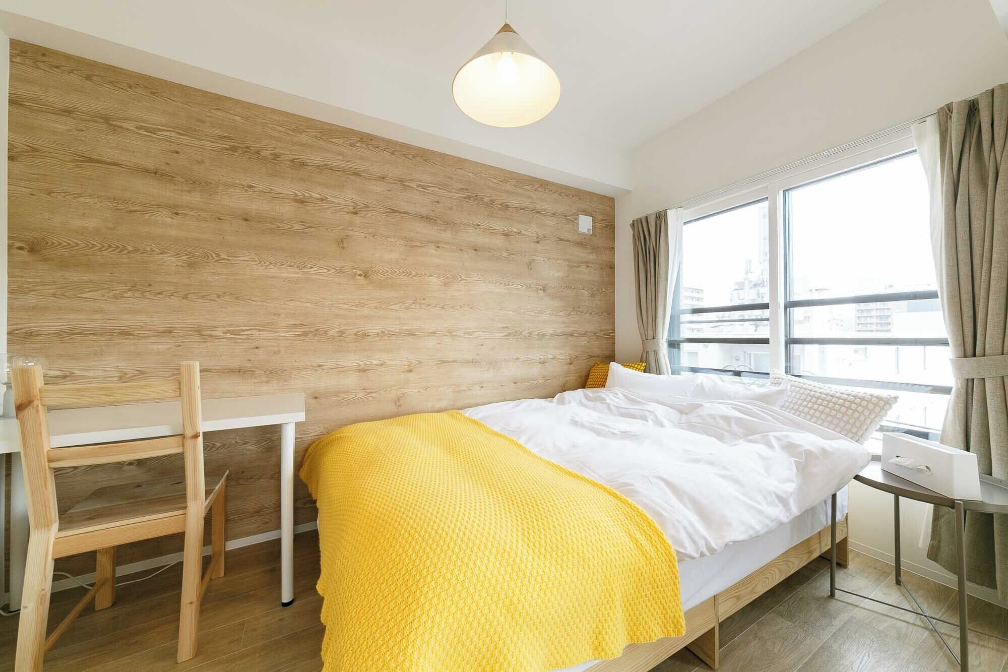 Un dormitorio con pared de madera, una cama con ropa blanca y un plaid amarillo, un escritorio y una ventana con cortinas.