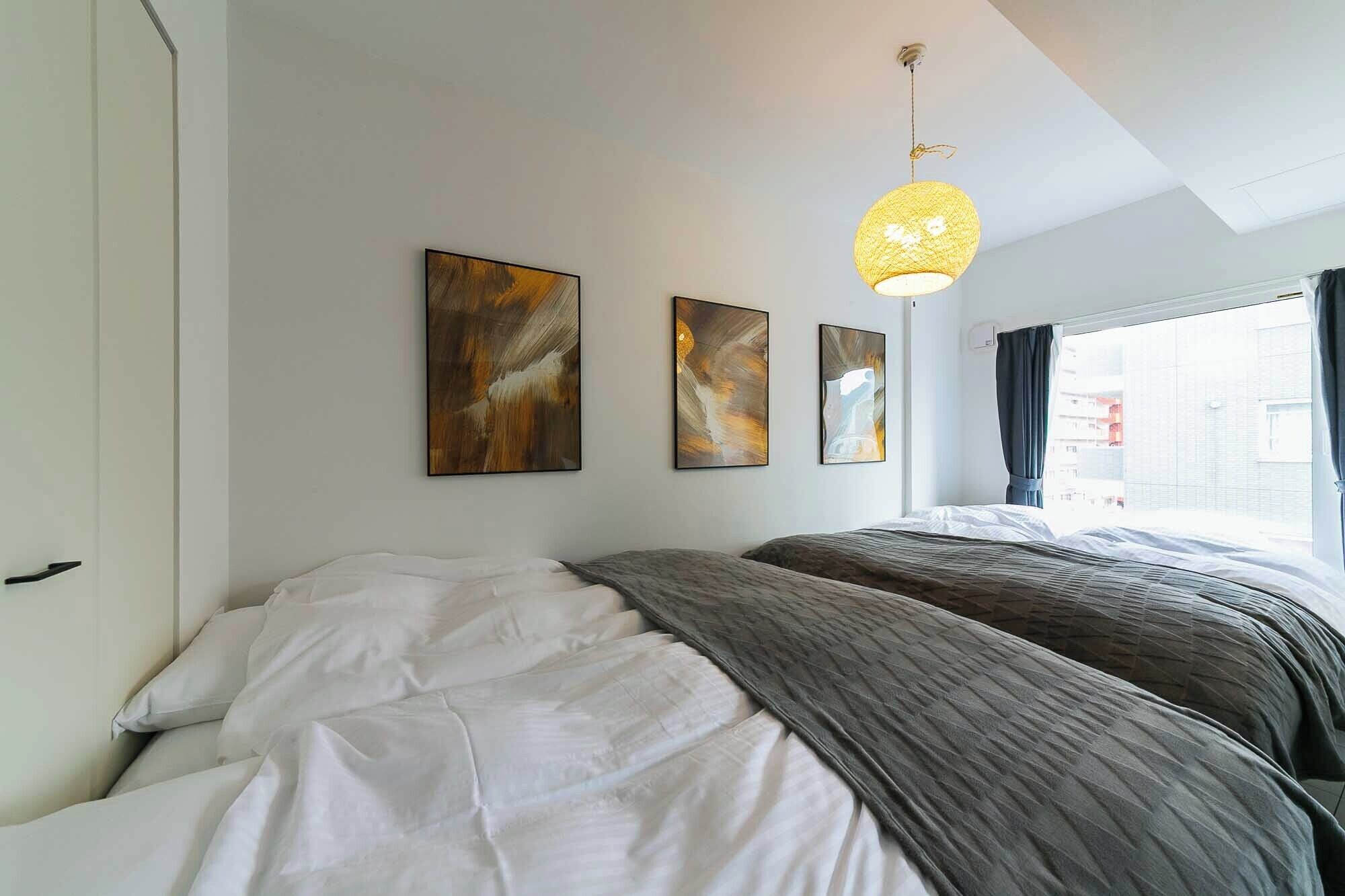 A bedroom with a bed, two wall-mounted abstract paintings, a hanging light fixture, and a window with curtains.