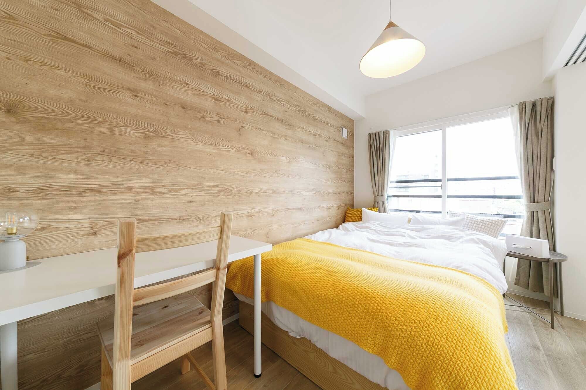 A modern hotel room with a wooden accent wall, a bed with a yellow blanket, a desk with a chair, and a large window offering a view.