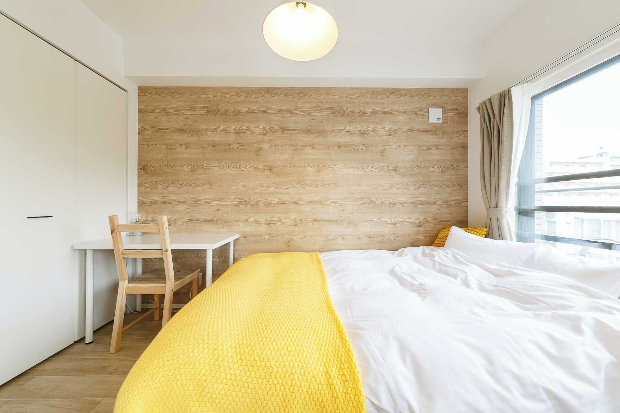 A hotel room with a wooden accent wall, a bed with white and yellow bedding, a desk with a chair, and a window with curtains.