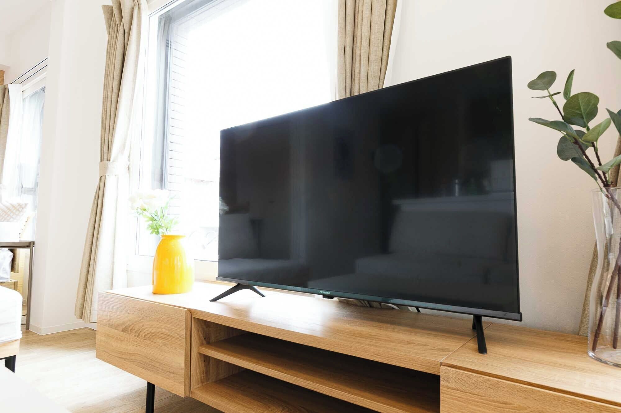 Una TV a schermo piatto su un mobiletto in legno in una stanza con finestra, tende e una pianta in vaso.