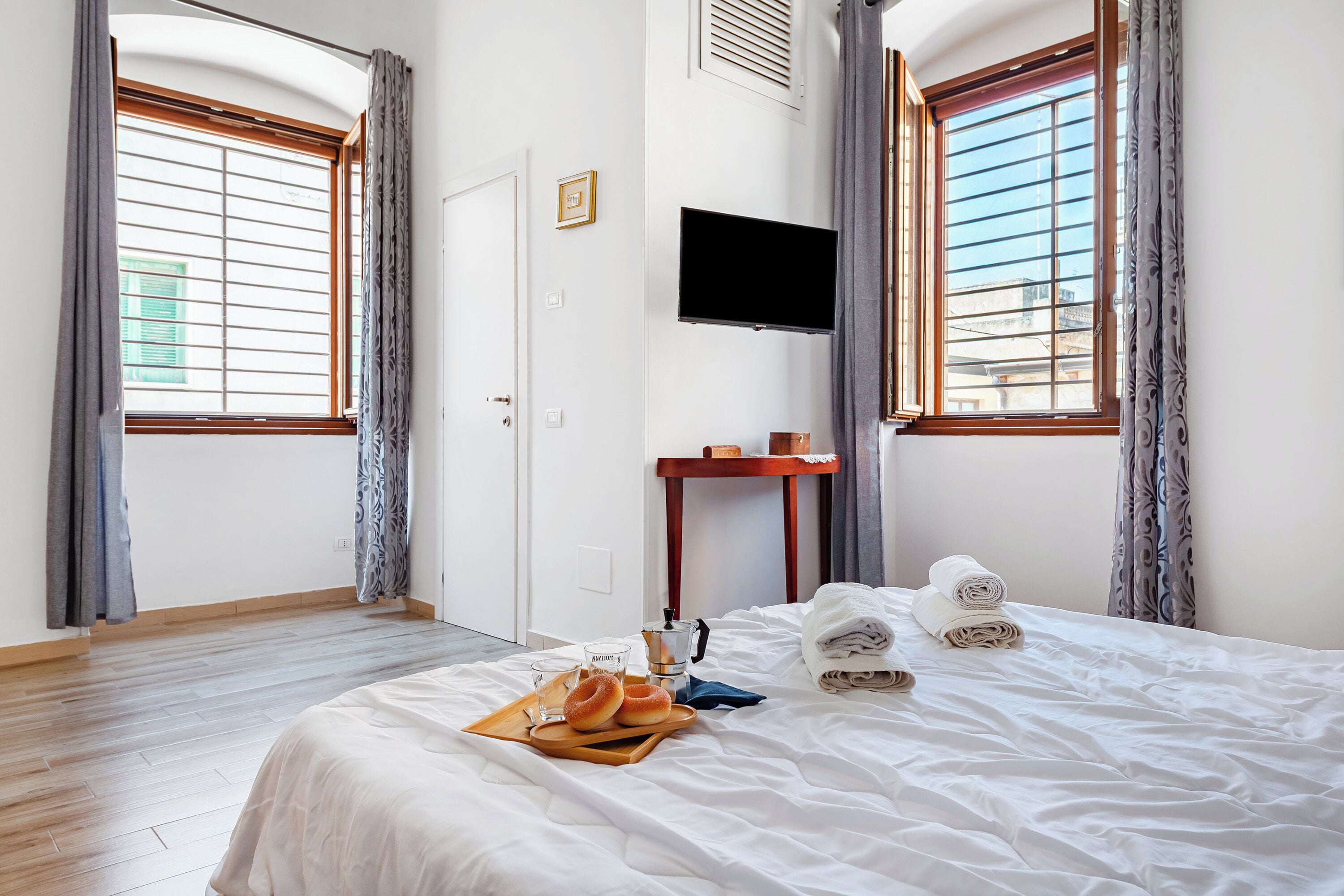 A hotel room with a bed, a flat-screen TV mounted on the wall, a small wooden table, and a window with curtains.