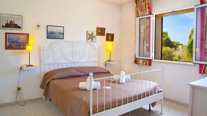A neatly made bed with a white metal frame, a brown bedspread, and two white towels. There are two wall-mounted lamps, a window with curtains, and several framed pictures on the wall. - Eurovillage Apartment with Pool & Terrace, Porto Cesareo, Italy (Porto Cesareo)
