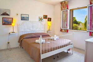 A neatly made bed with a white metal frame, a brown bedspread, and two white towels. There are two wall-mounted lamps, a window with curtains, and several framed pictures on the wall. - Eurovillage Apartment with Pool & Terrace, Porto Cesareo, Italy (Porto Cesareo)