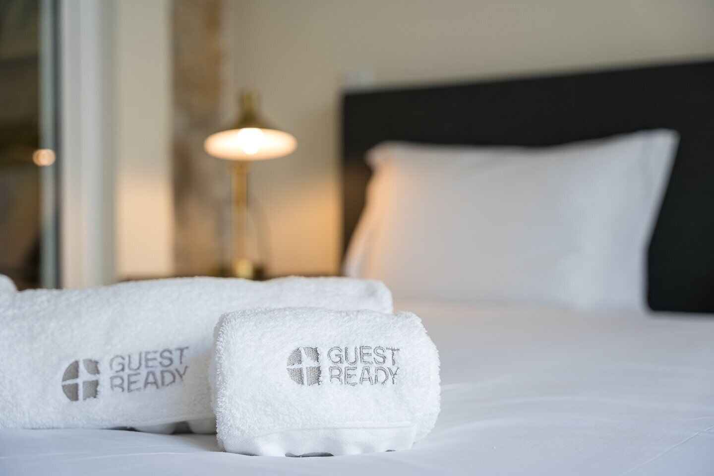 Een net opgemaakt bed met twee witte handdoeken erop, elk met de tekst 'GUEST READY' gedrukt.
