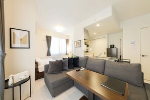 A modern hotel room with a grey sofa, a wooden table, a bed with white linens, a small side table with a bottle, and a framed picture on the wall. - EFFECT Kikusui (Sapporo)
