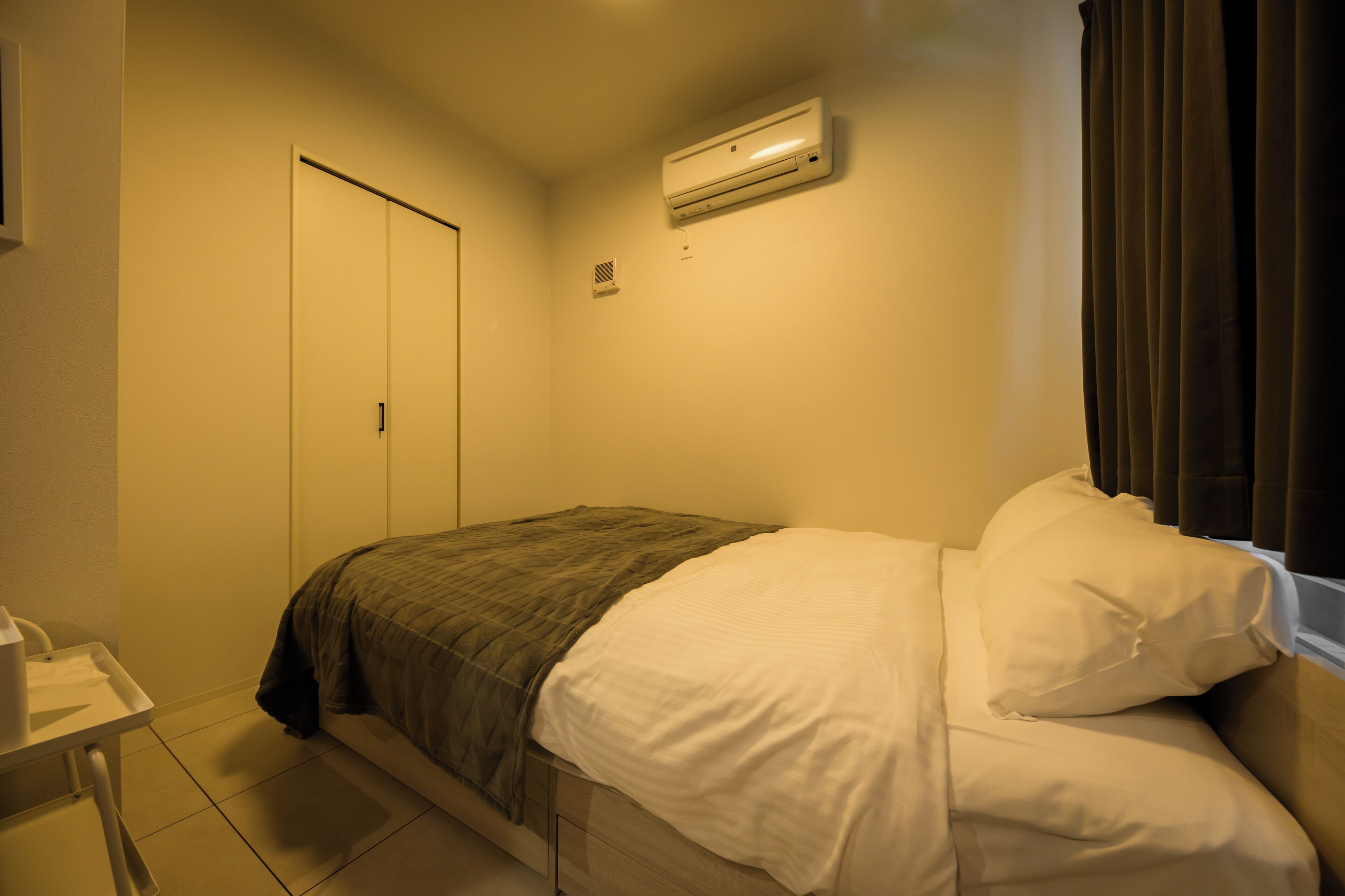 A hotel room with a bed, a bedside table, a wardrobe, and an air conditioning unit.
