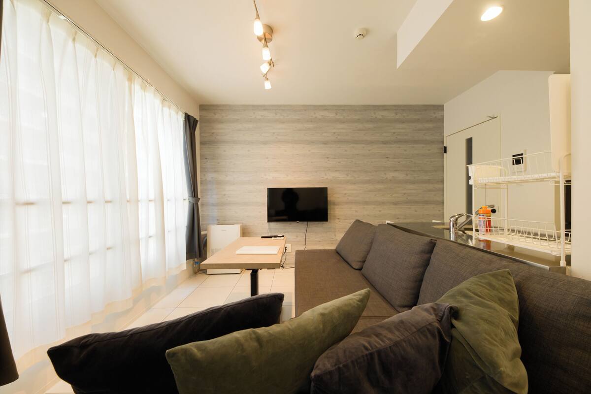 Una sala de estar moderna con sofá, televisor de pantalla plana, mesa de comedor y zona de cocina con lavaplatos y estanterías.