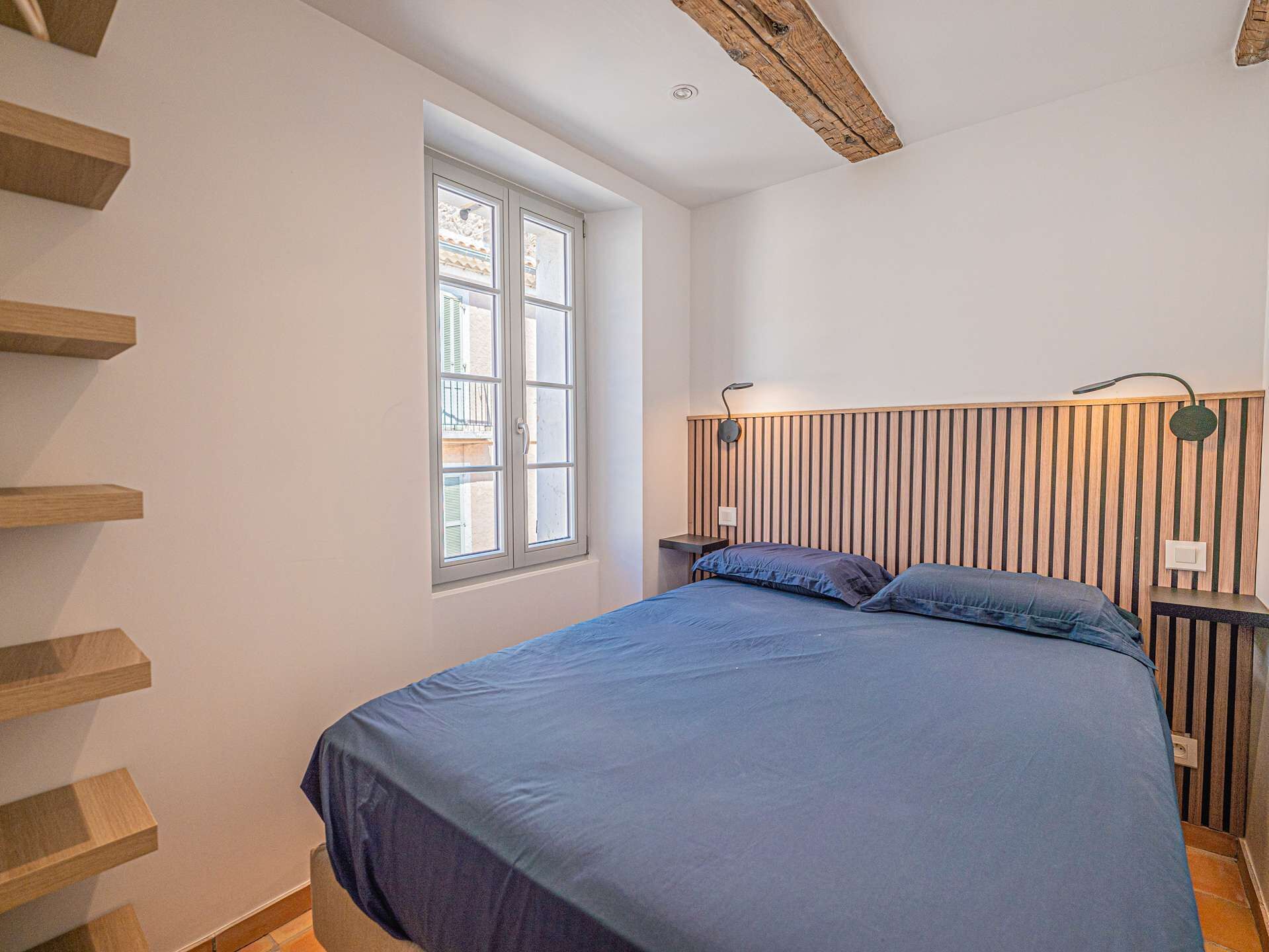 Ein Schlafzimmer mit Bett, Holz-Kopfstütze, Nachttischen, einem Fenster und einer Holztreppe.
