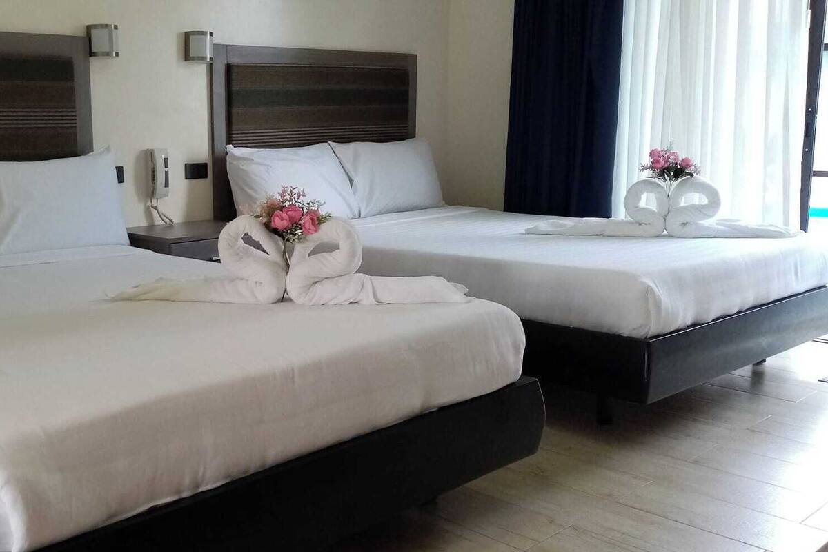 Habitación de hotel con dos camas, cada una con sábanas y almohadas blancas. Hay toallas decorativas sobre las camas y un jarrón con flores en la mesita de noche.