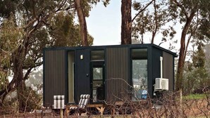 A modern, dark-colored cabin with a glass door, a chair, and a small table outside. - Churinga Orchards by Tiny Away (Lenswood)